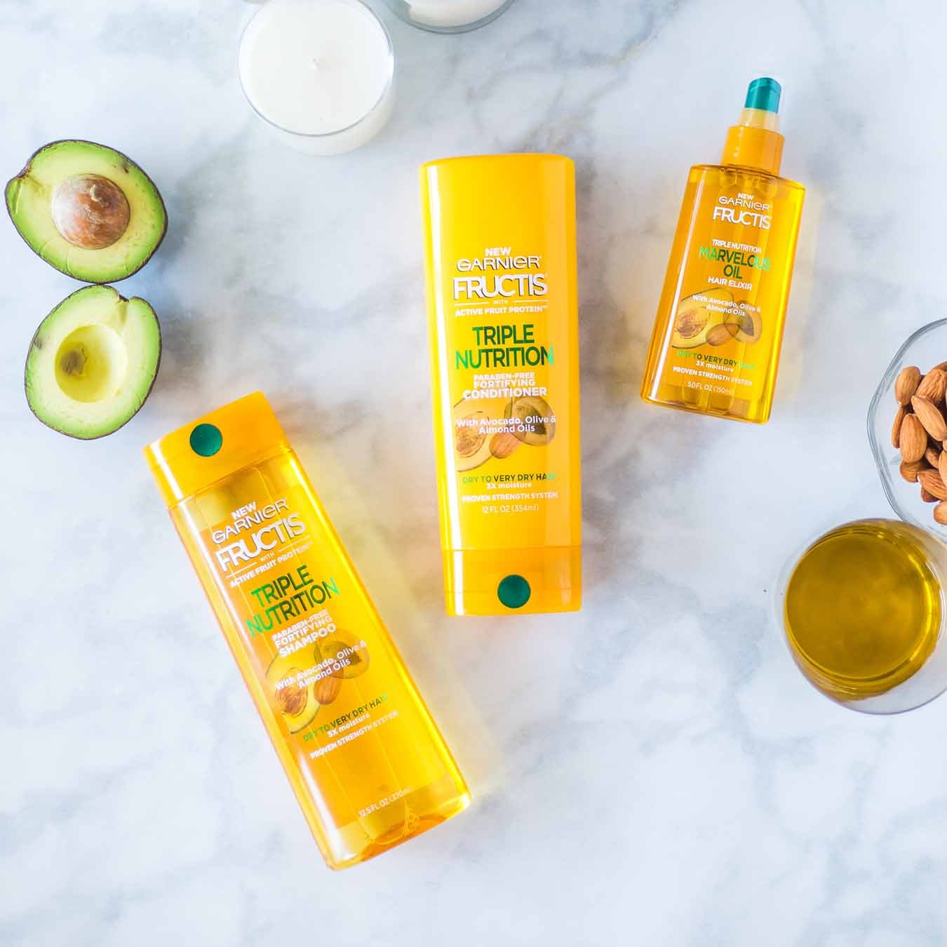 Garnier Fructis Triple Nutrition Shampoo, Fructis Triple Nutrition Conditioner, and Fructis Marvelous Oil on white marble next to a halved avocado, two white candles, a glass of oil, and a bowl of almonds.